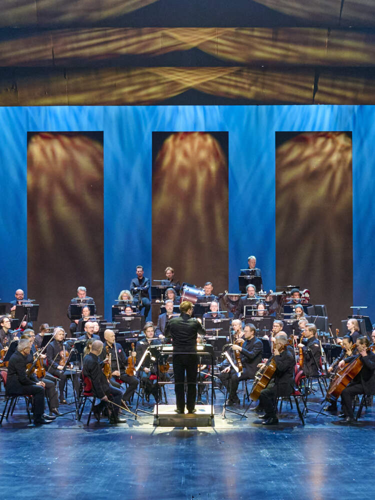 En stor orkester och dirigent är på en upplyst teaterscen.