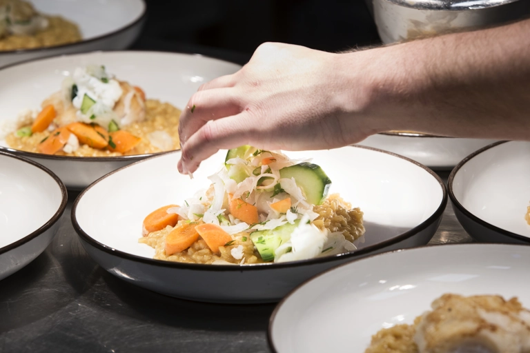 En hand garnerar en rätt som står på tallrik under serveringslampor på en restaurang. Liknande rätter är uppställda i närheten.