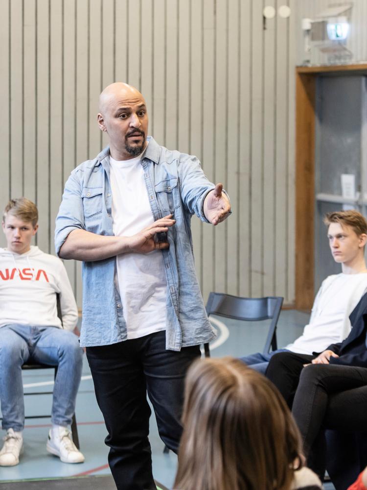 En operasångare står och pratar i ett skolgym, bland en grupp elever som sitter på stolar runt honom.