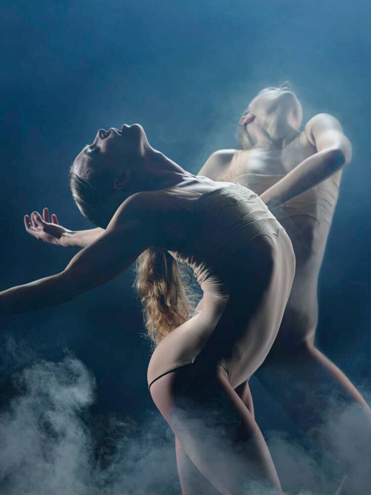 Two dancers stand under white stage lighting in smoke, the dancer in the foreground stands with arms outstretched behind her.