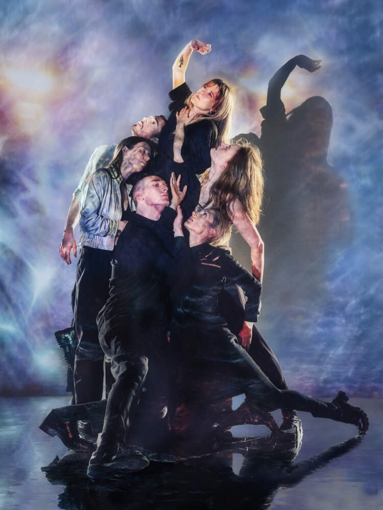 Sex dansare står tätt runt Anna som höjs över dem med en arm sträckt uppåt. Ljus projiceras över gruppen och fonden bakom.