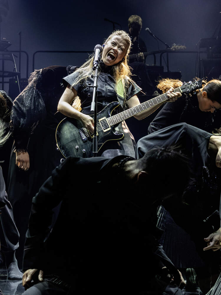 En sångare under scenljus med en elgitarr vrålar i en mikrofon. Runt henne står flera svartklädda dansare med böjda huvuden.