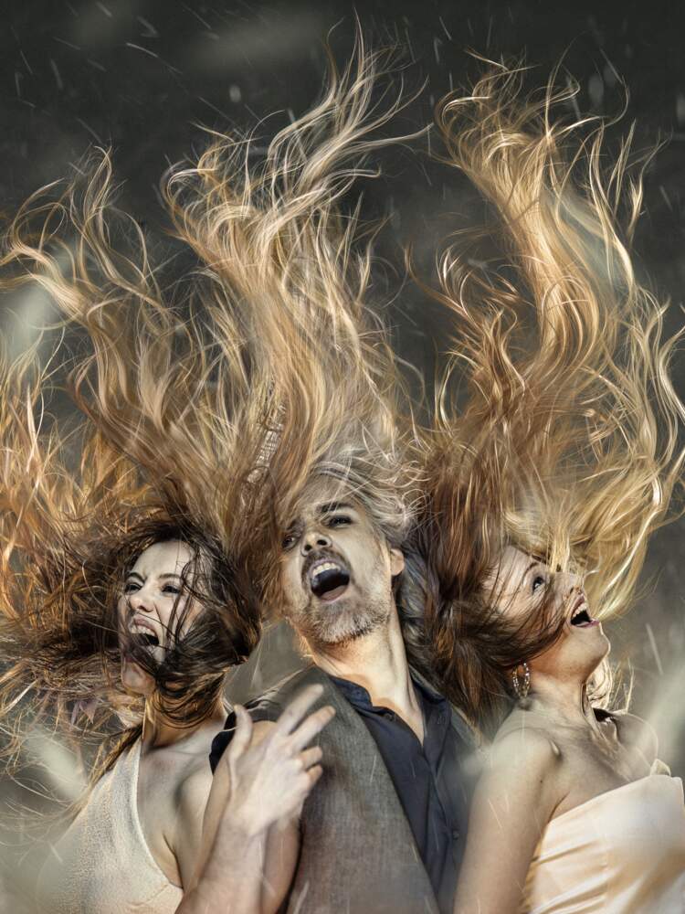 Tre körsångare står rygg mot rygg och glädeligen sjunger högt, medan deras långa hår kraftigt blåses uppåt underifrån.