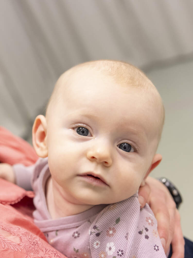 En ung bebis som hålls av sin mamma tittar rakt in i kameran. Hennes mamma tittar på henne och ler.