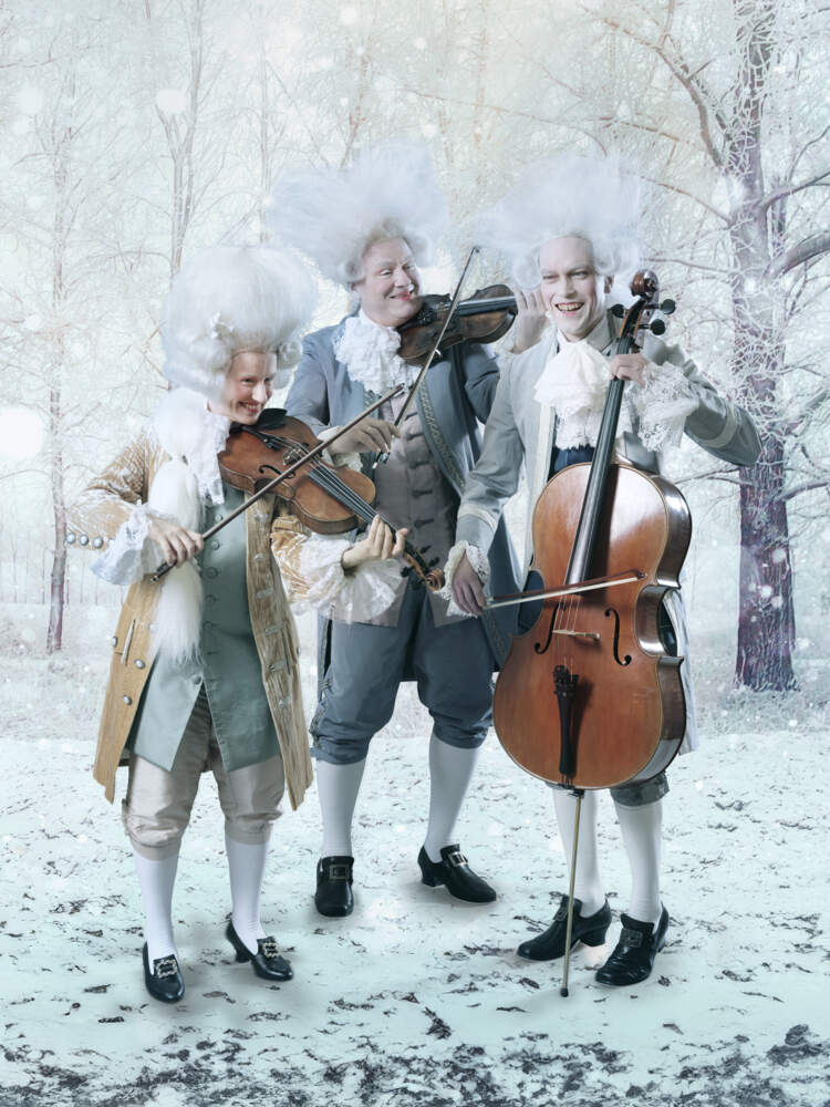Tre stråkmusiker i barocka kläder och stora vita peruker står och spelar sina instrument på ett snöigt fält omringad av träd.
