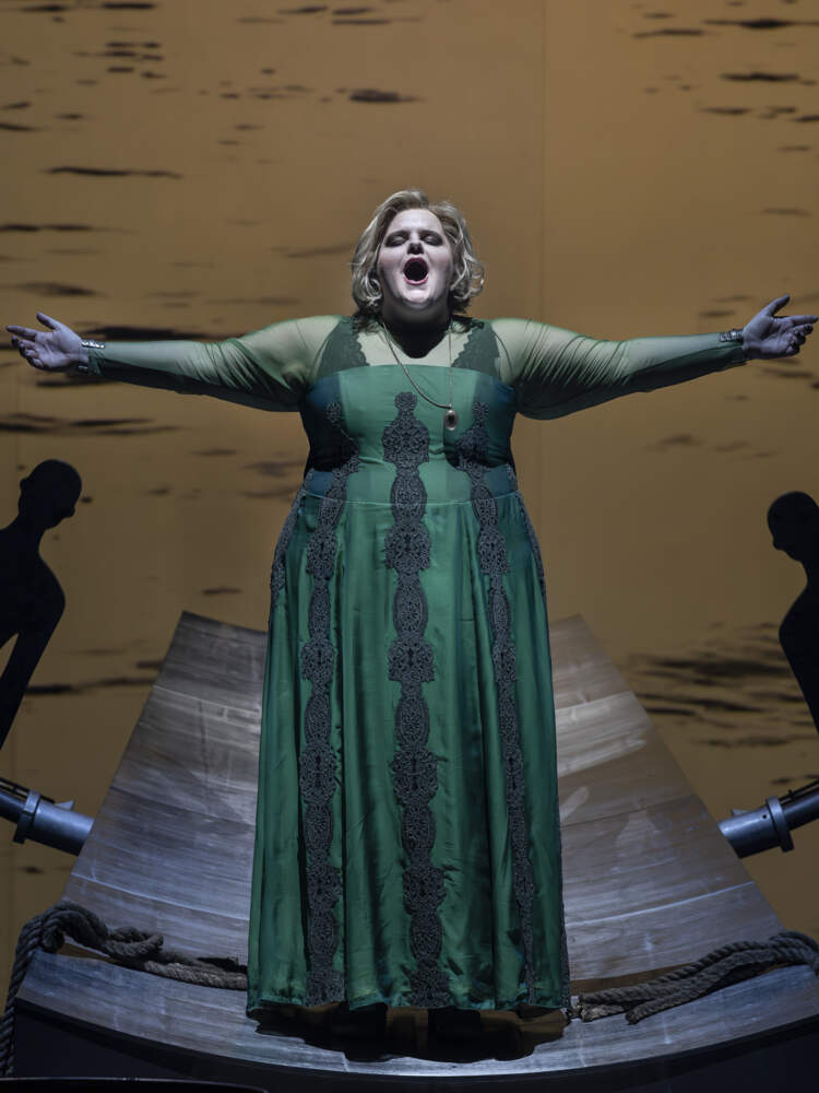 Senta står och sjunger med armarna utsträckta på en plattform framför en stor portal gjord av mänskliga silhuetter.