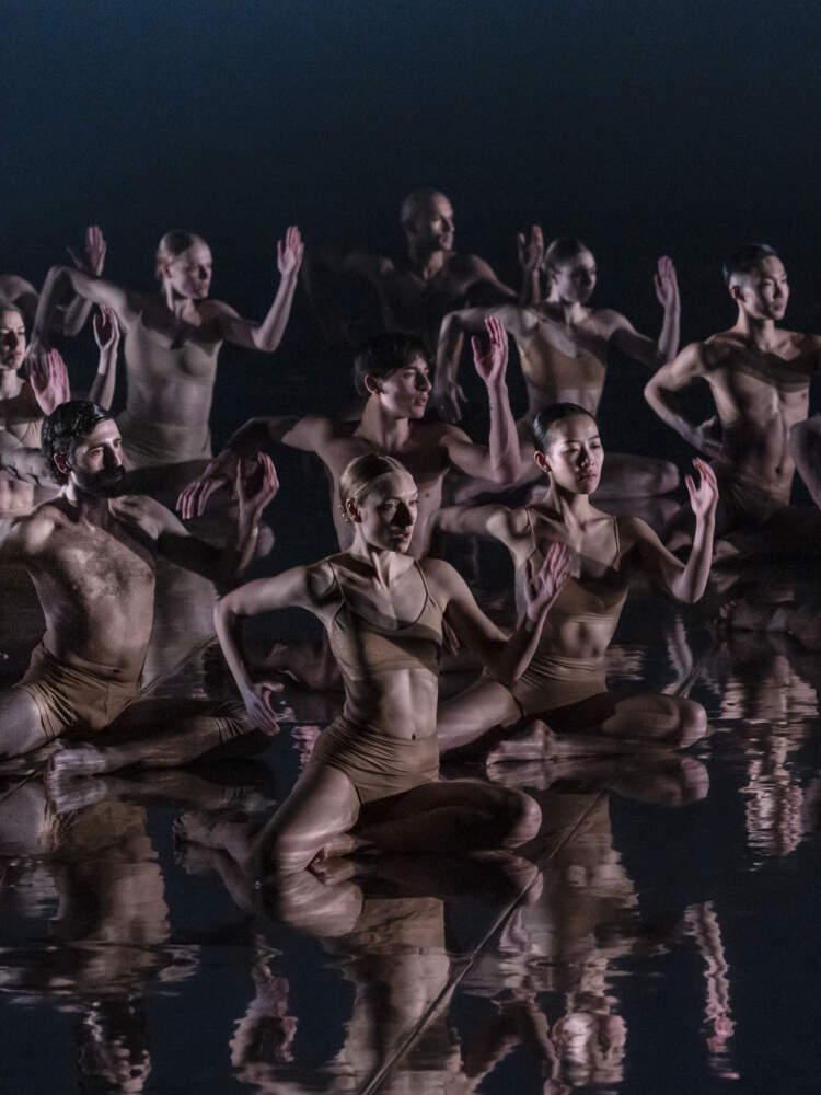 En grupp dansare sitter på ett spegelgolv, alla vända åt höger. De håller i deras armar utsträckta som om de springer.