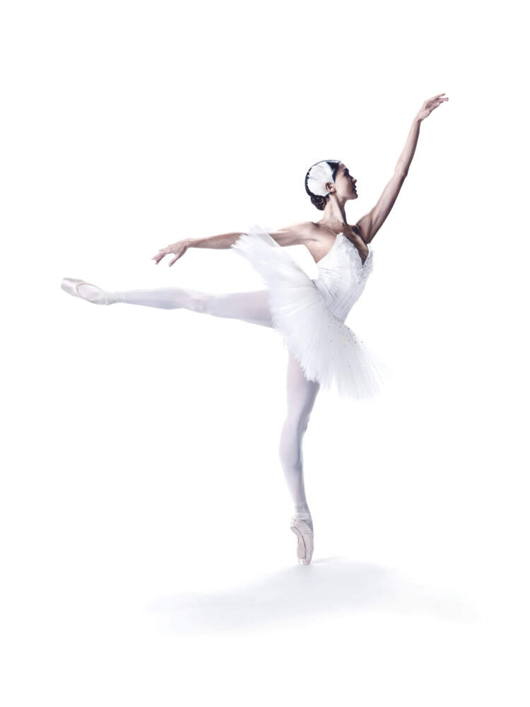 En ballerina i vit tutu balanserar på ena tån med en arm sträckt fram och ett ben utsträckt bakom sig.