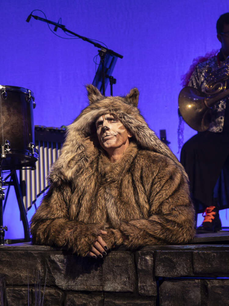 Sandvargen sitter bakom en låg stenmur och funderar. Bakom honom i mörkret sitter två musiker som spelar deras instrument.