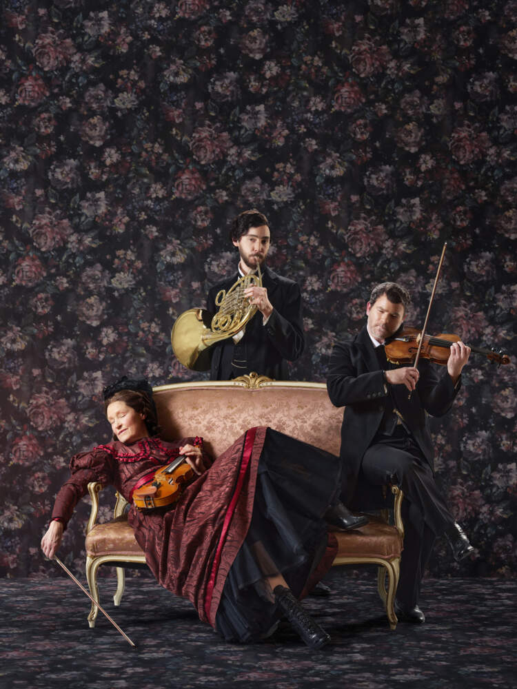 En violinist ligger med slutna ögon på en schäslong i ett tapetserat rum, medan en annan violinist samt hornist spelar.