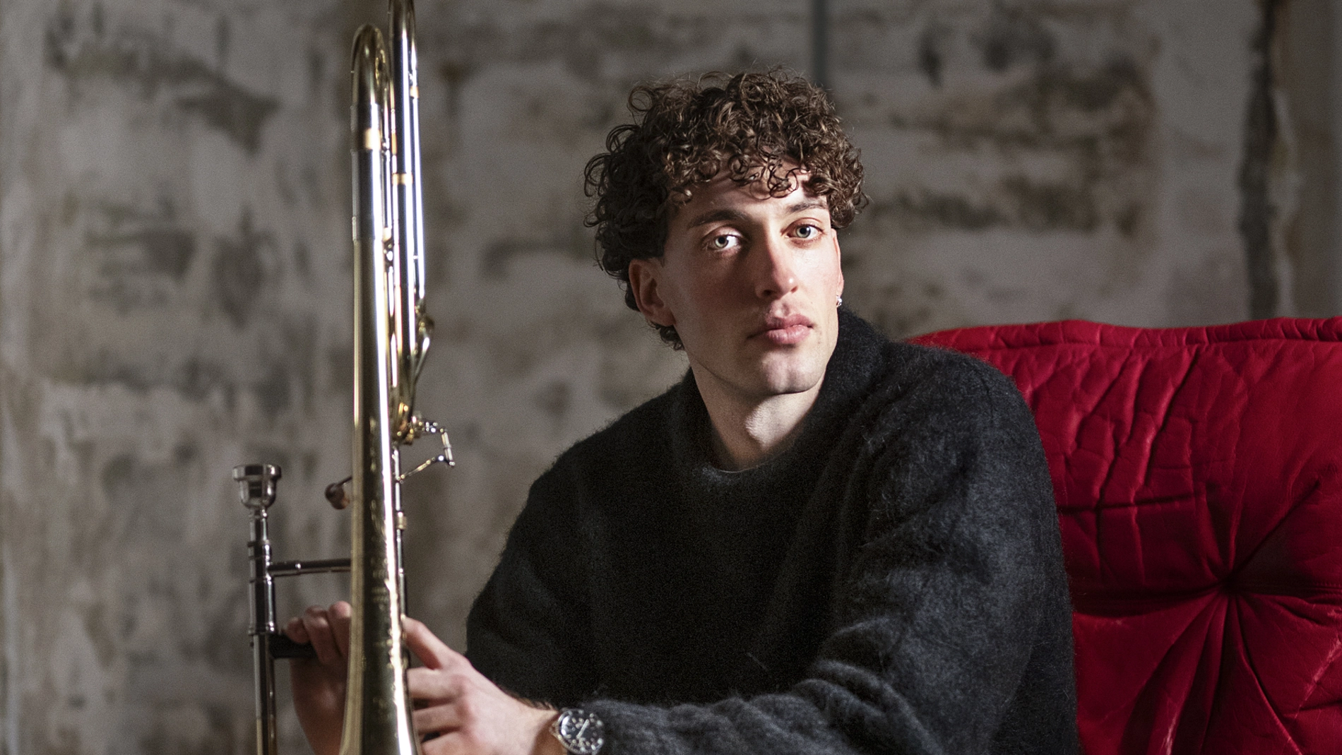 En trombonist sitter i en röd läderfåtölj och håller sitt instrument. Han är barfota och är upplyst från vänster sida.