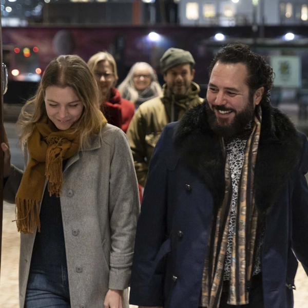 Ett leende par i vinterjackor går in i en teaterfoajé genom glasdörrar. Bakom dem går tre personer till mot entrén.