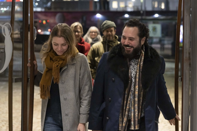 Ett leende par i vinterjackor går in i en teaterfoajé genom glasdörrar. Bakom dem går tre personer till mot entrén.